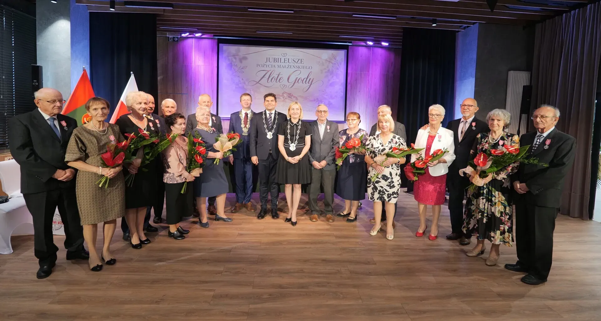 Grupa jubilatów pozuje razem z bukietami czerwonych róż na tle banera z napisem Złote Gody.