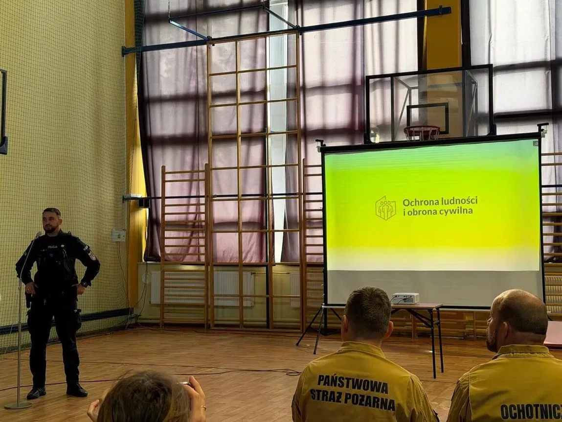 Mężczyzna w ciemnym mundurze mówi do mikrofonu w sali gimnastycznej; za nim ekran z napisem „Ochrona ludności i obrona cywilna”, po prawej strażacy w żółtych kurtkach.