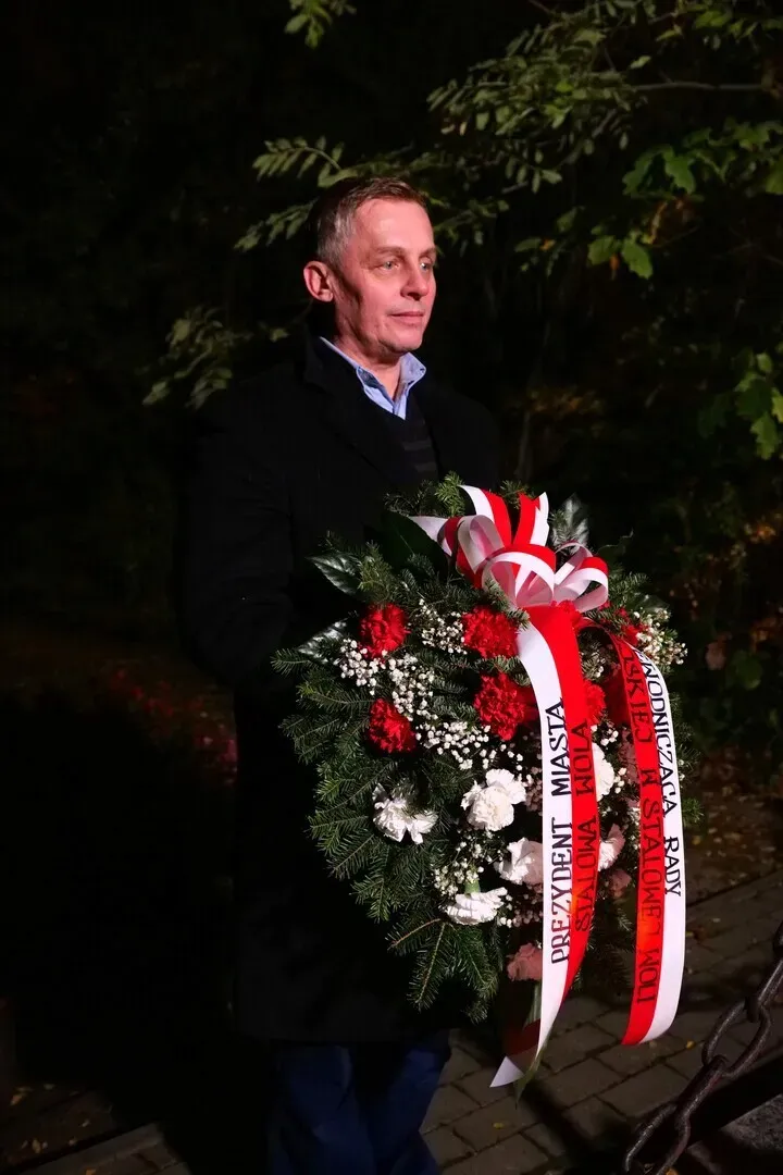 Mężczyzna w ciemnym płaszczu trzyma wieniec z biało-czerwonymi wstęgami, stojąc w nocnym otoczeniu drzew.